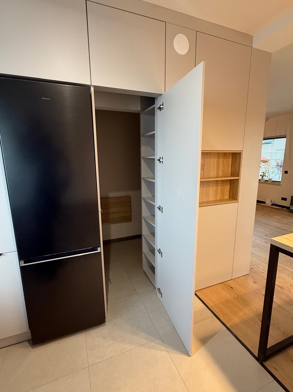 Nowoczesna zabudowa kuchenna z czarną lodówką i otwartą szafką z półkami, widok na wnętrze szafki i fragment blatu. Minimalistyczny design, jasne kolory.