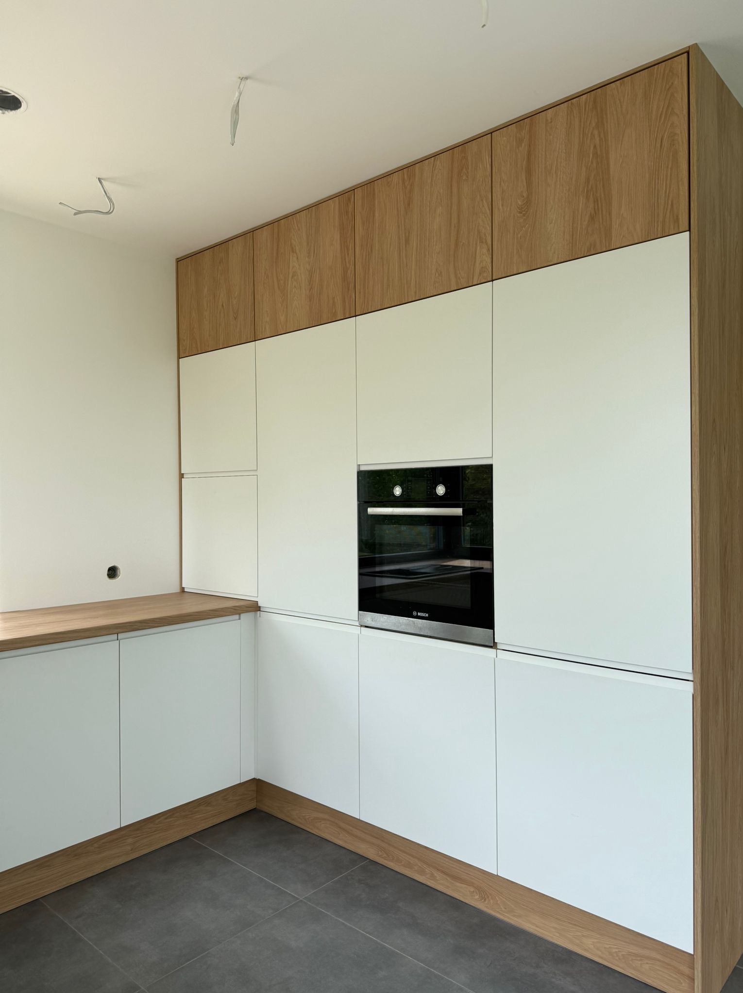 Nowoczesna kuchnia na wymiar: białe fronty szafek z drewnianymi akcentami, wbudowany piekarnik Bosch, szare płytki podłogowe. Minimalistyczny design.