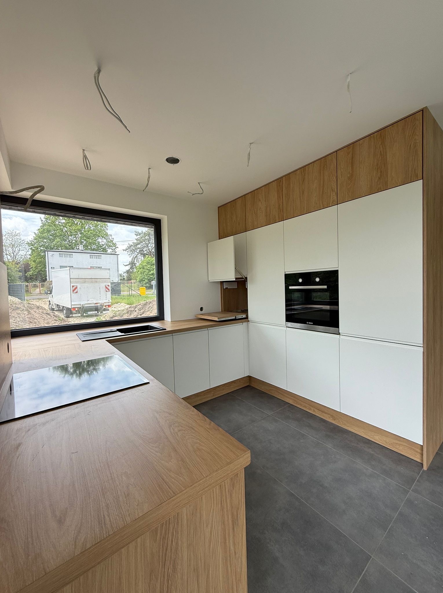 Nowoczesna kuchnia w trakcie montażu: białe fronty, drewniane akcenty, płyta indukcyjna. Widoczne kable zwisające z sufitu i okno z widokiem na plac budowy.