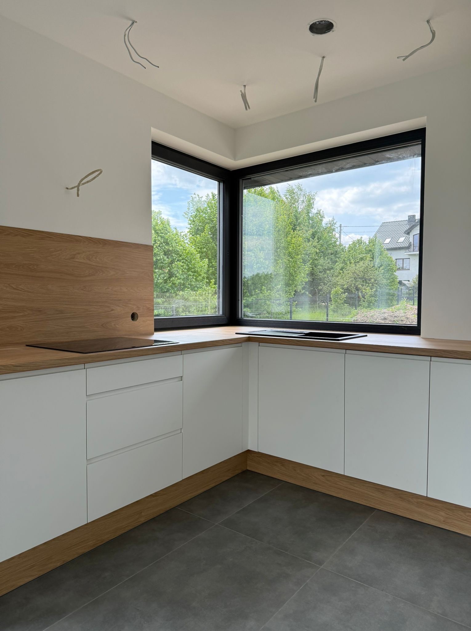 Jasna kuchnia z drewnianym blatem i białymi szafkami, duże okno z widokiem na zieleń. W tle widać wystające kable elektryczne.