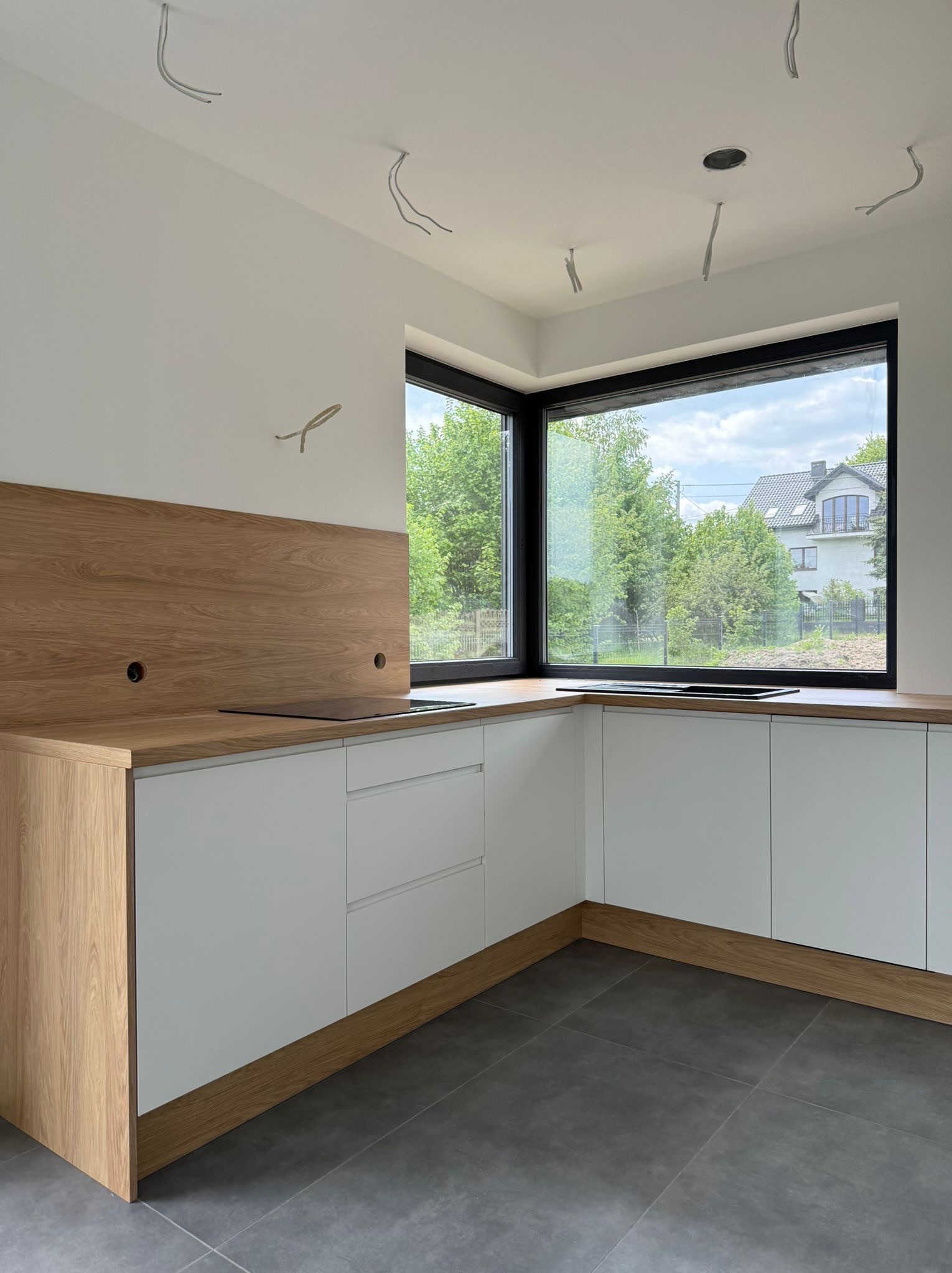 Nowoczesna kuchnia w trakcie montażu: białe fronty, drewniany blat i panel, duże okno z widokiem na zieleń. Widoczne kable elektryczne.