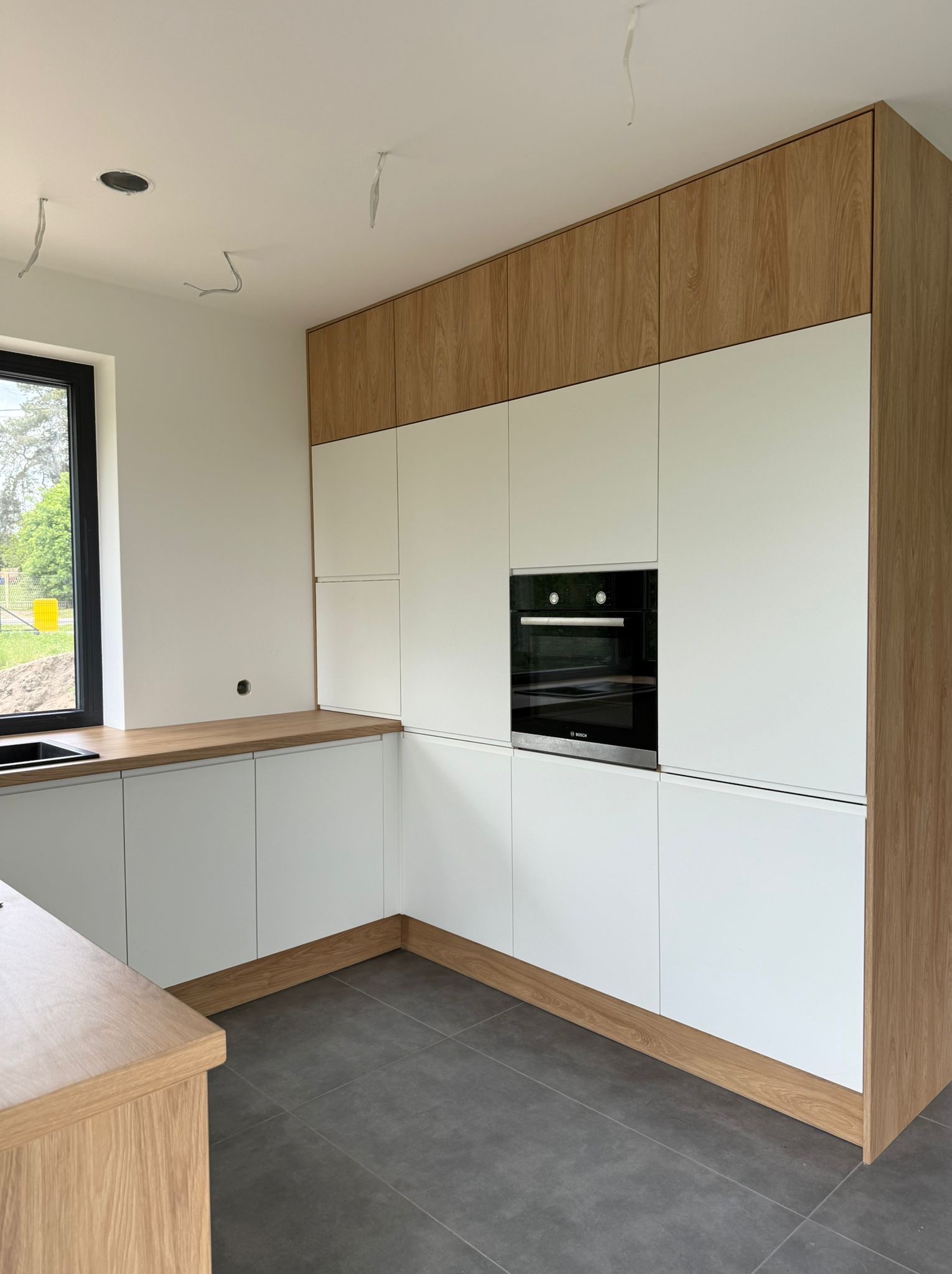 Nowoczesna kuchnia z białymi frontami i drewnianymi akcentami. Widoczny piekarnik w zabudowie i szare płytki podłogowe. W tle okno z widokiem na zieleń.