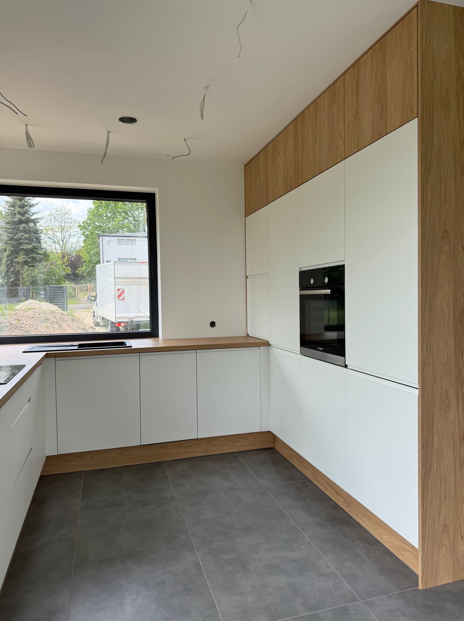 Nowoczesna kuchnia w bieli i drewnie: minimalistyczne fronty, drewniane akcenty, szare płytki podłogowe. Widok na zewnątrz przez okno.