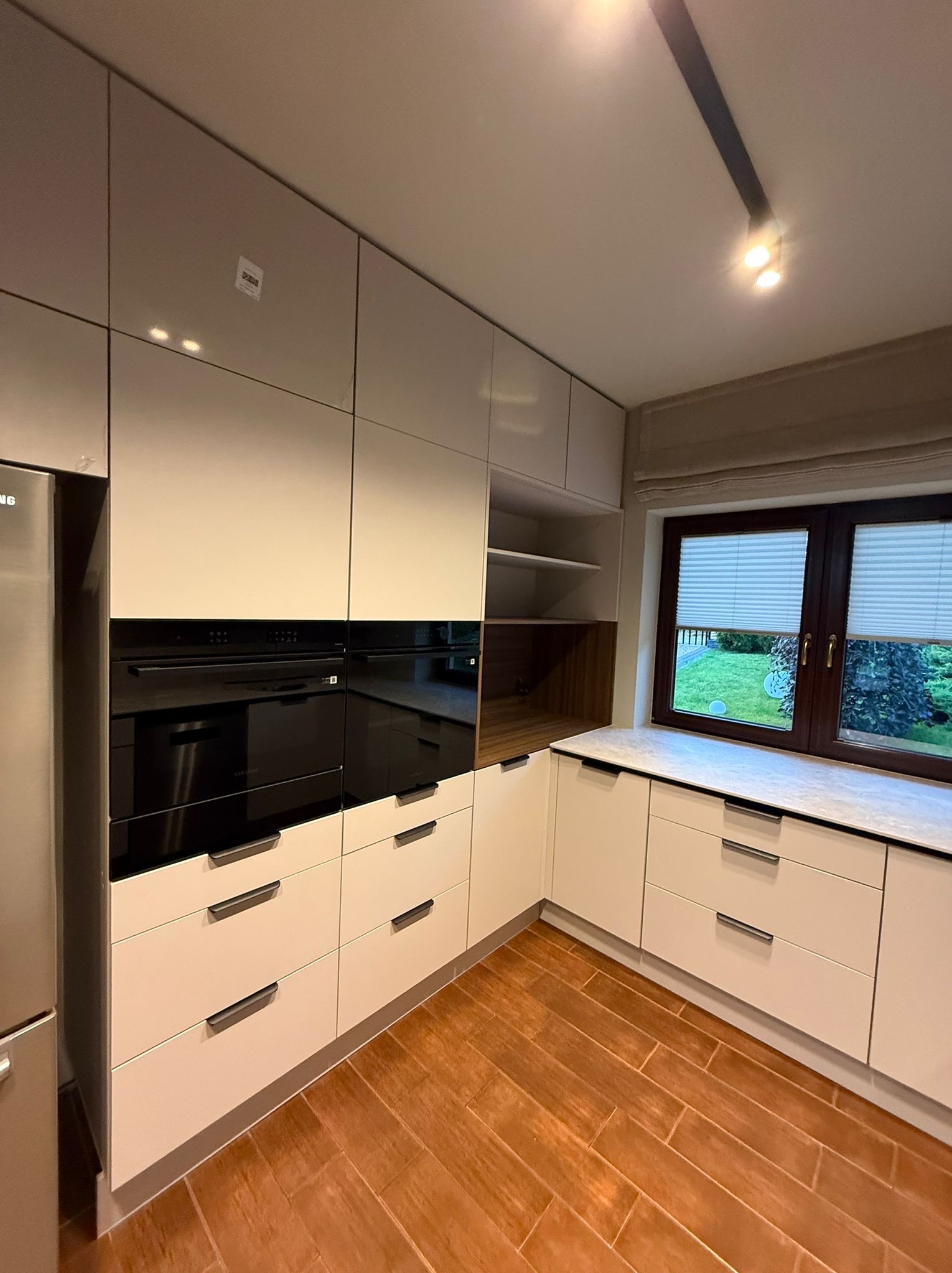 Jasna kuchnia z zabudową pod sufit, czarnymi piekarnikami i drewnianą podłogą. Okno z roletami w tle. Minimalistyczne uchwyty szafek.