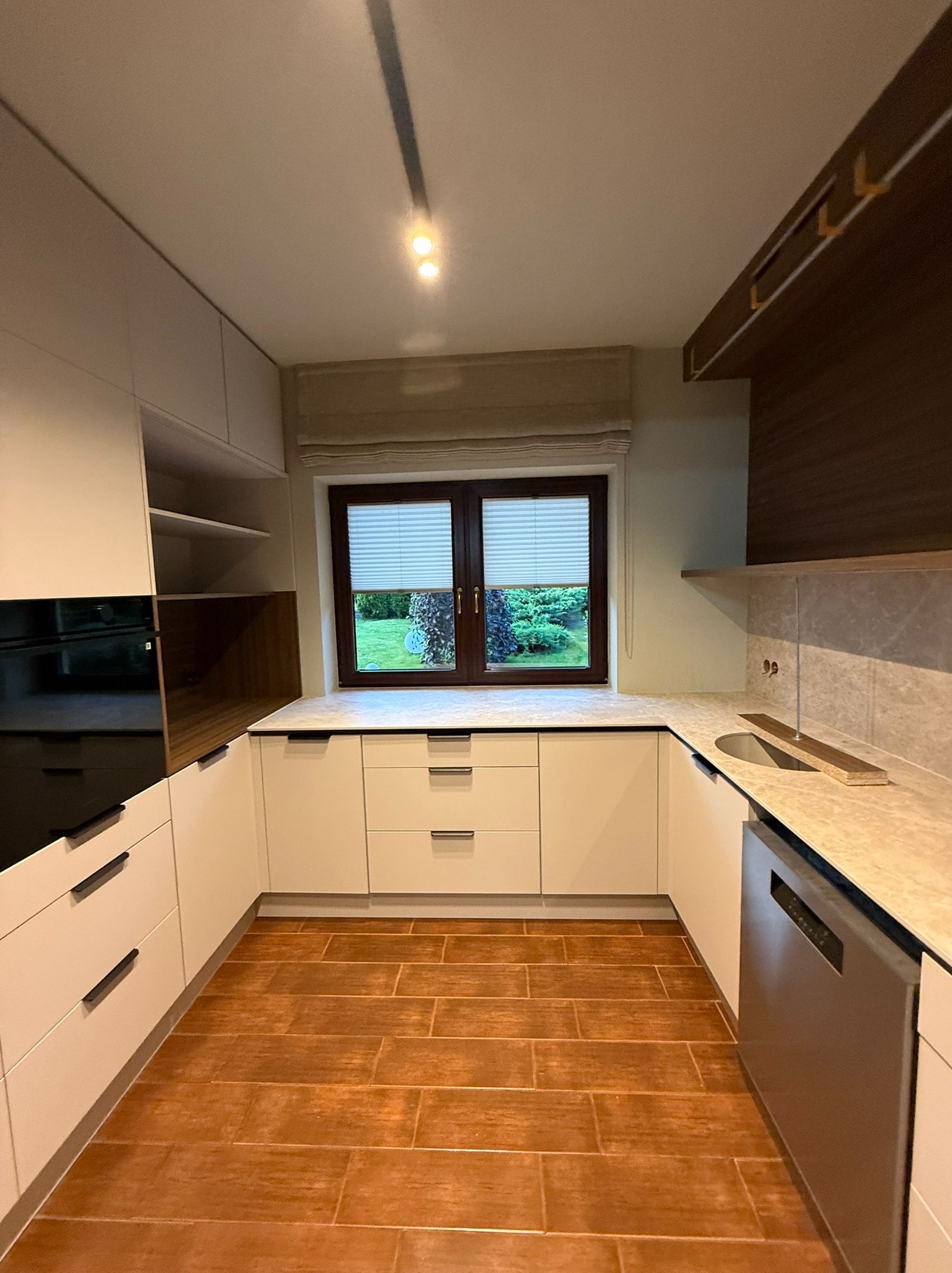 Nowoczesna kuchnia w kształcie litery U z białymi frontami, drewnianymi akcentami i oknem z widokiem na zieleń. Brązowa podłoga imitująca drewno. Minimalistyczny design.