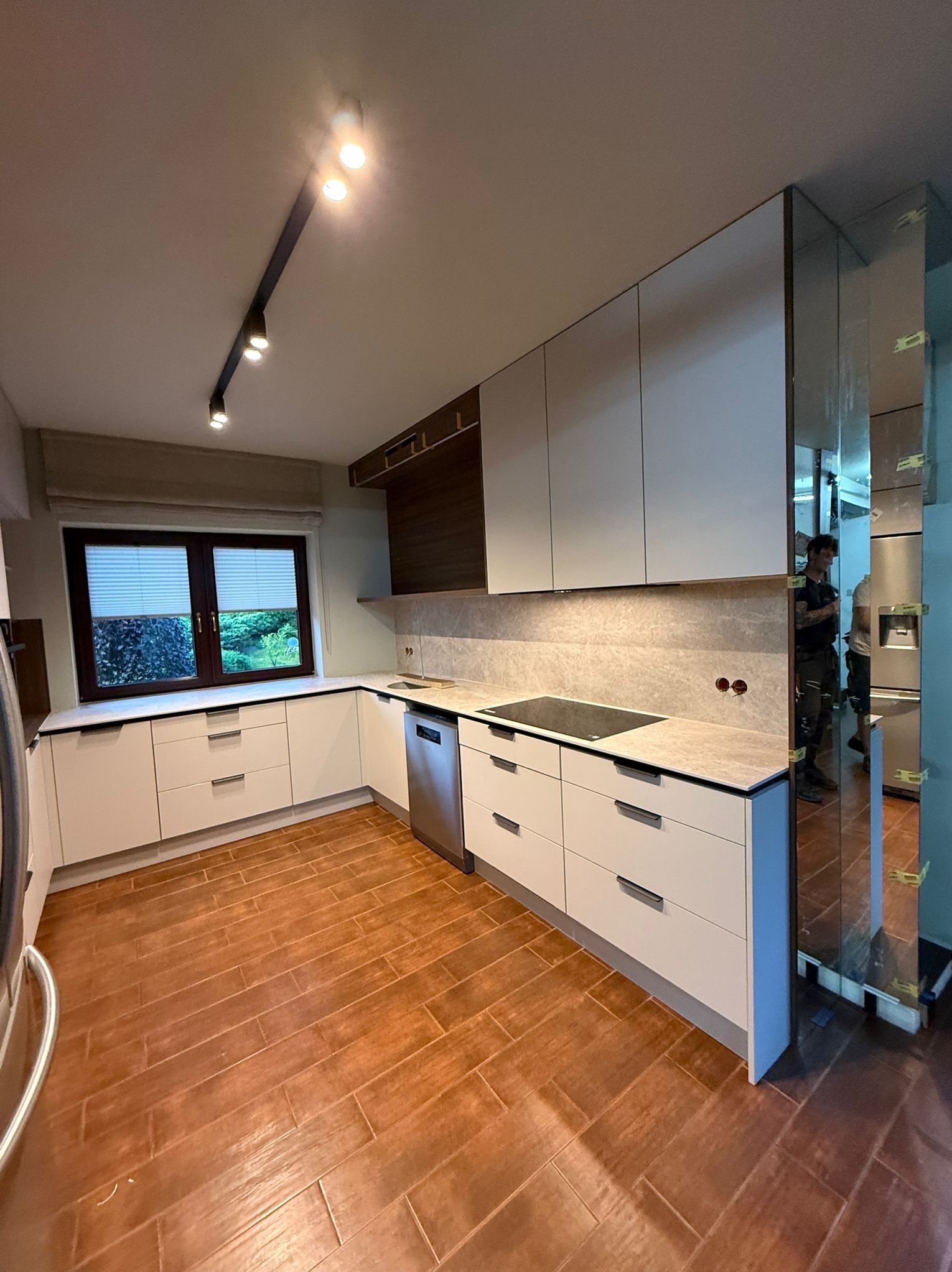 Jasna kuchnia z drewnianymi akcentami i oświetleniem punktowym. Widoczne szafki, blat, okno z żaluzjami i fragment podłogi z płytek imitujących drewno. W lustrze odbija się osoba.