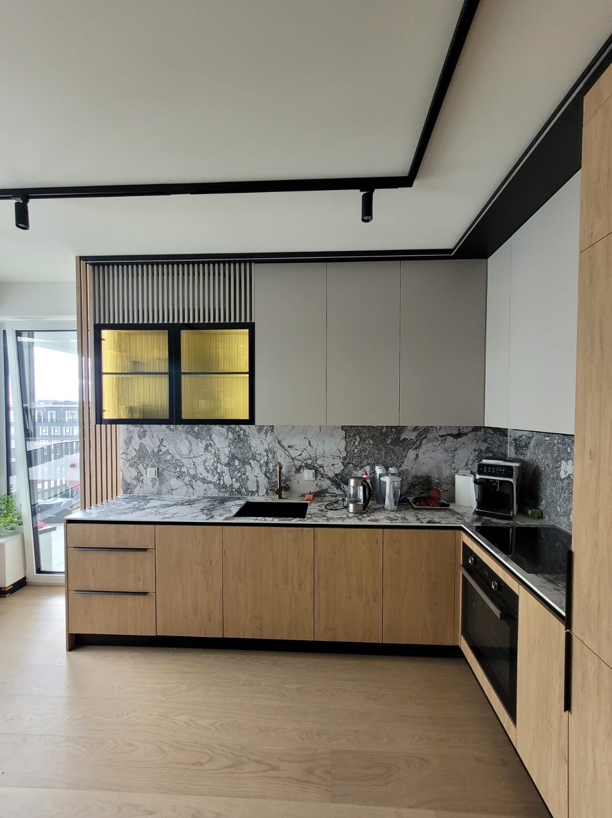 Nowoczesna kuchnia z drewnianymi frontami, marmurowym blatem i złotymi akcentami. Widok na miasto przez okno. Oświetlenie punktowe na suficie.