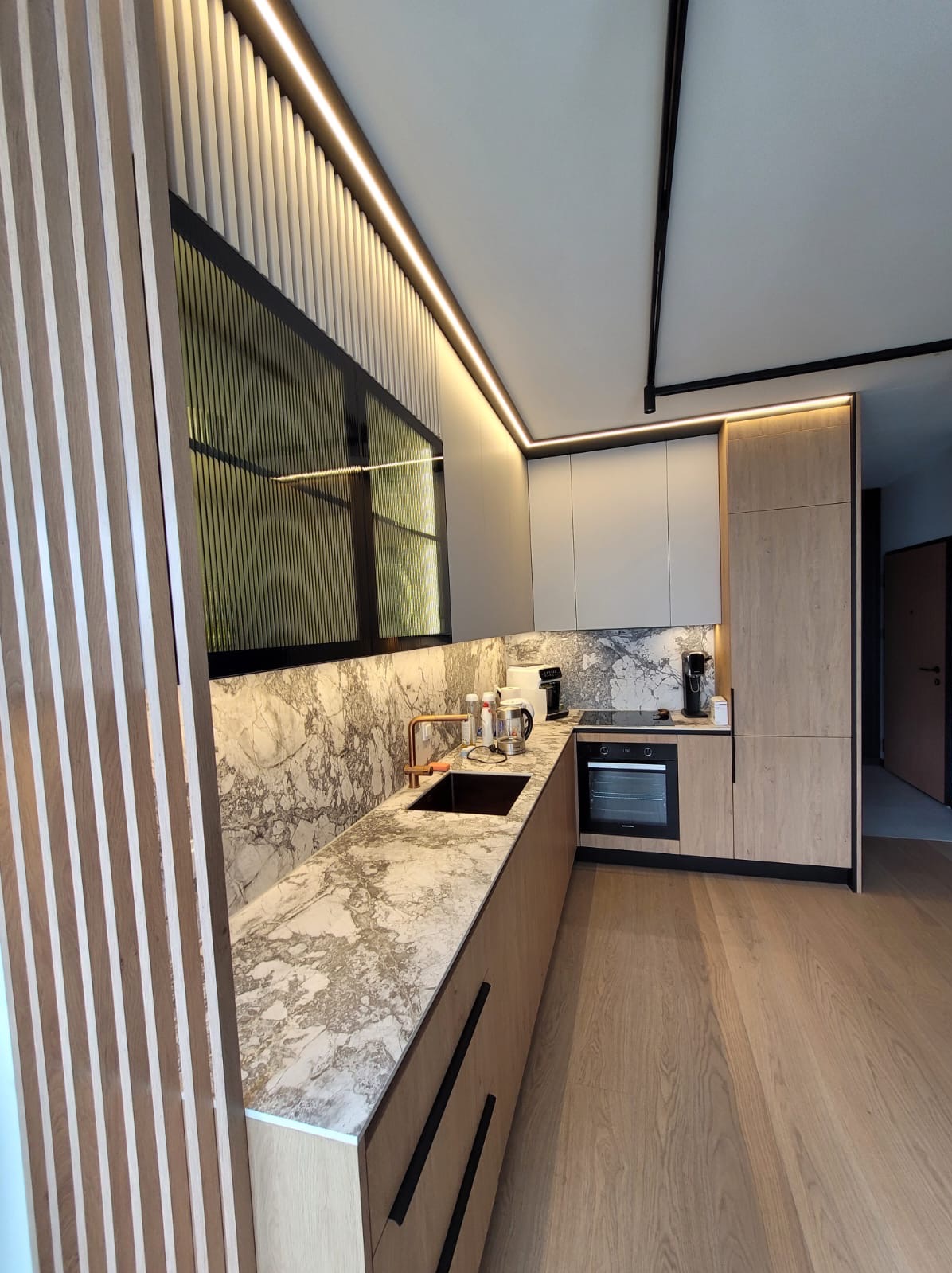 Nowoczesna kuchnia z marmurowym blatem i drewnianymi frontami, oświetlenie LED podkreśla unikalny design. Widoczne detale: czarna armatura i pionowe lamele dekoracyjne.