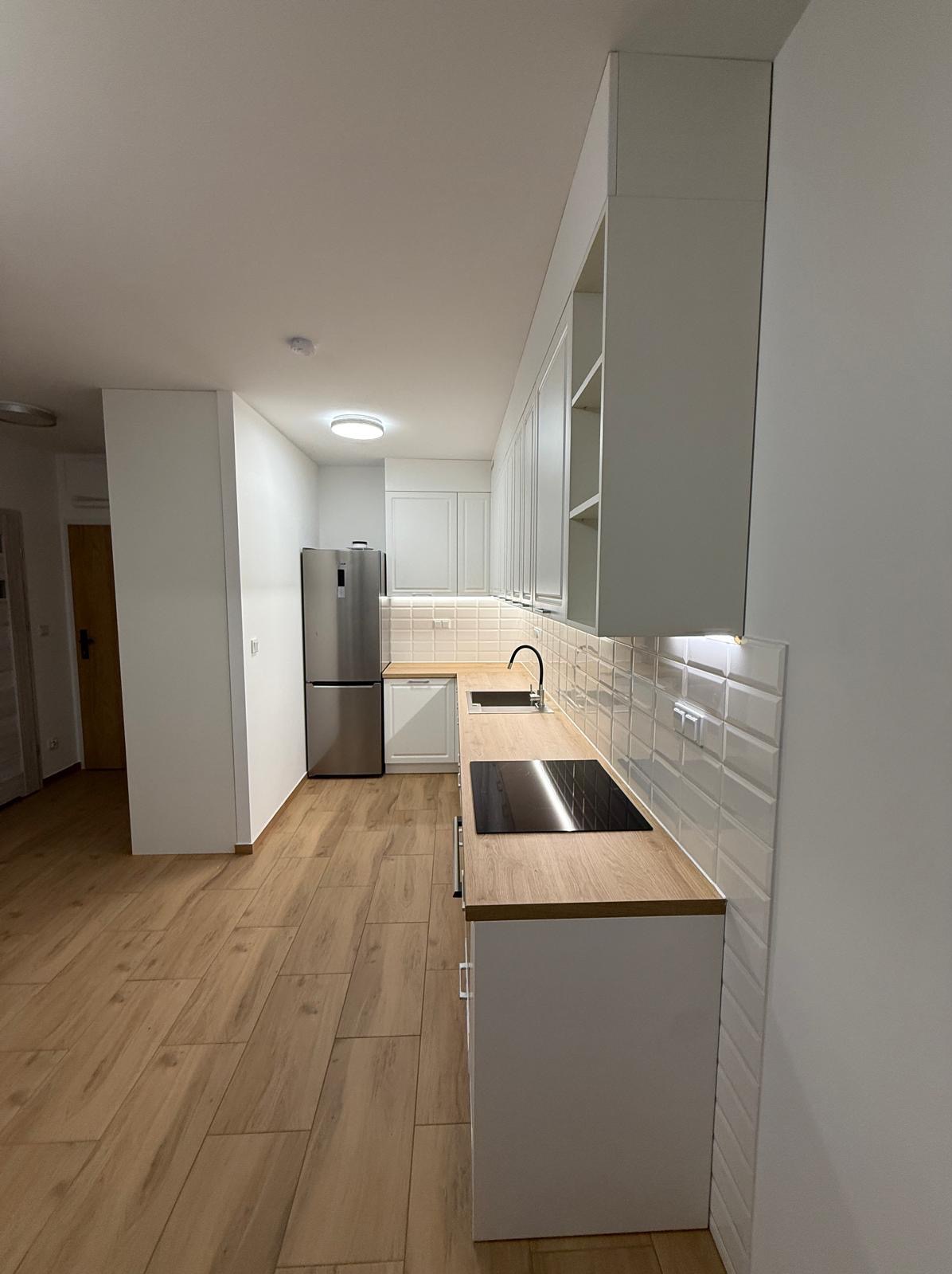 Jasna kuchnia z drewnianym blatem i białymi szafkami. Widoczna płyta indukcyjna, zlew i lodówka. Podłoga wyłożona panelami imitującymi drewno.