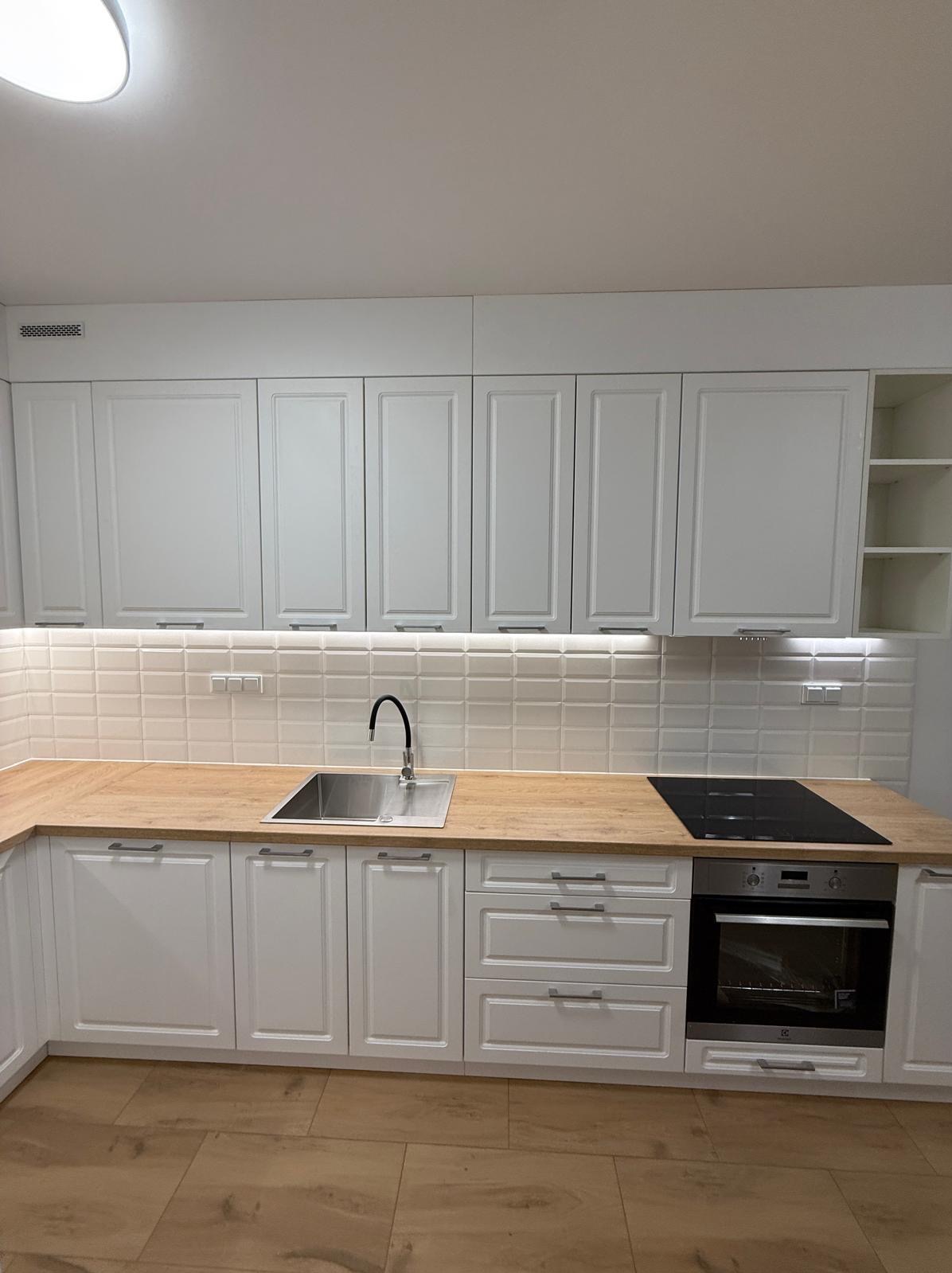 Biała kuchnia z drewnianym blatem i płytkami w stylu retro. Nowoczesne oświetlenie podszafkowe podkreśla detale. Minimalistyczny design z elementami klasycznymi.