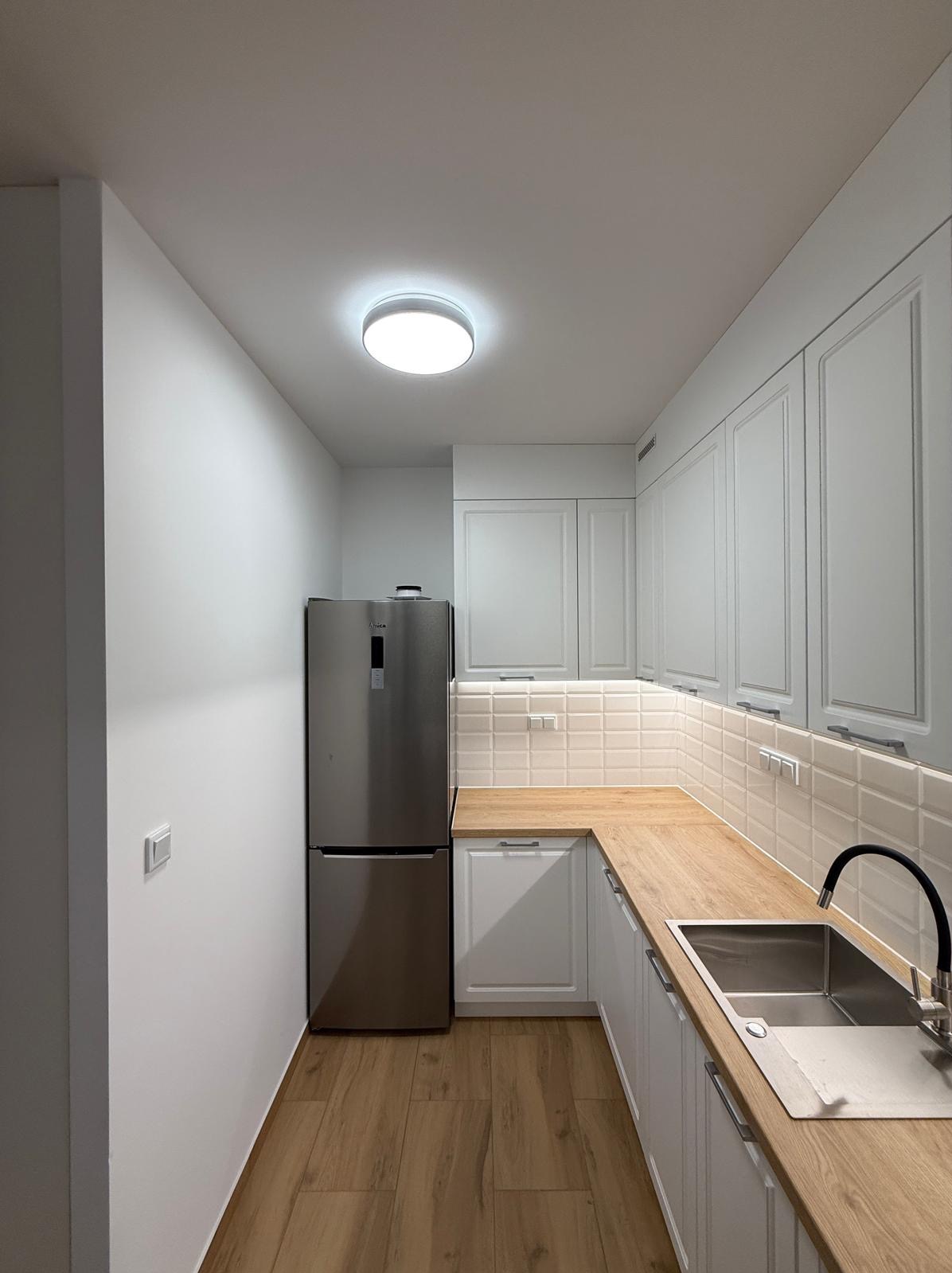 Jasna kuchnia z drewnianym blatem i białymi szafkami. Uwagę przykuwa srebrna lodówka Amica i minimalistyczne oświetlenie sufitowe. Nowoczesny design.