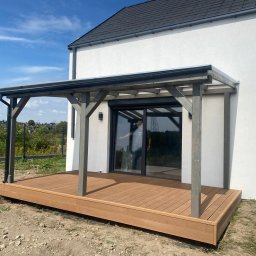 TARAS OD ZARAZ - Drewniany taras kompozytowy z szarą pergolą przy białym domu. Widoczny fragment ogrodu i niebieskie niebo. Osoba w odbiciu szyby.