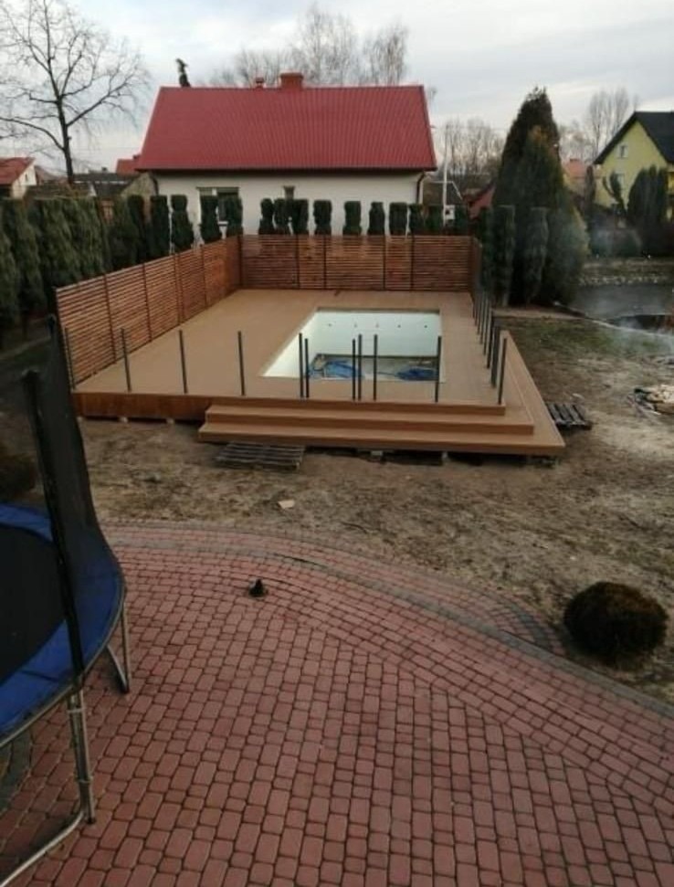 Kompozytowy taras z basenem w ogrodzie, otoczony drewnianym ogrodzeniem. Widok z góry na czerwoną kostkę brukową i trampolinę. Dom z czerwoną dachówką w tle.