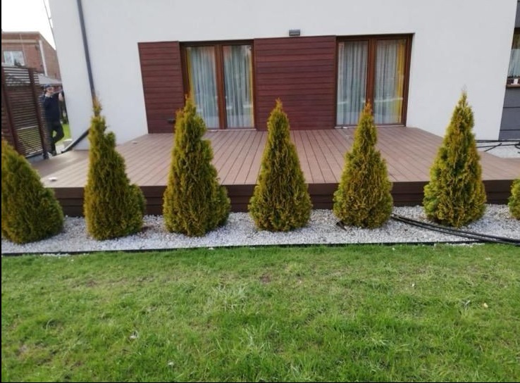 Nowoczesny taras z deski kompozytowej, obłożony ozdobnymi kamieniami i obsadzony tujami. Widoczny fragment elewacji budynku z brązowymi panelami.