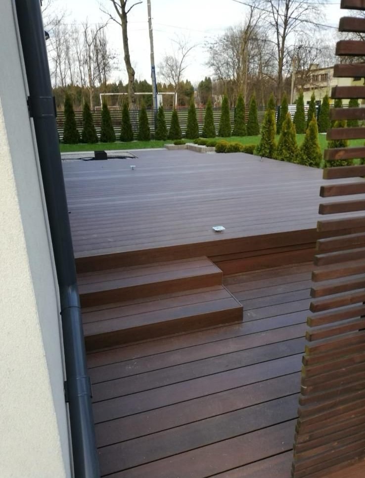 Nowoczesny taras kompozytowy z niskimi schodami i ażurową przesłoną, otoczony zielenią. Widok na zadbany ogród z tujami i boiskiem w tle.