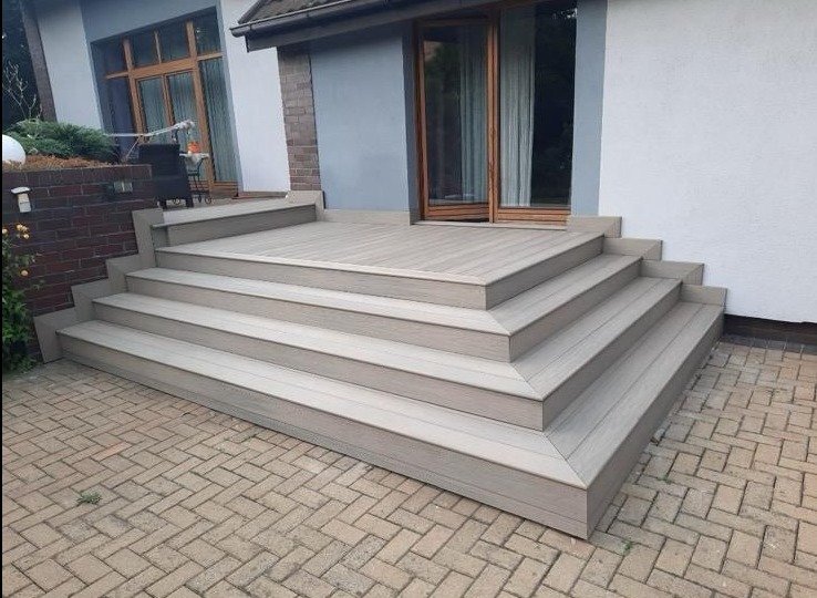 Jasny taras kompozytowy z czterema stopniami prowadzącymi do drzwi balkonowych. Widoczna kostka brukowa i fragment elewacji budynku.