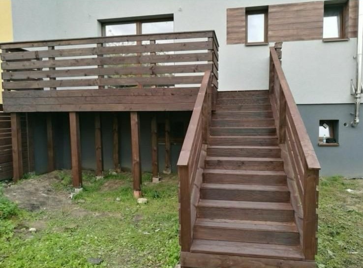 Drewniany taras z balustradą i schodami prowadzącymi do ogrodu, przy szarym budynku z drewnianymi elementami elewacji. Widok z dołu, trawa w tle.
