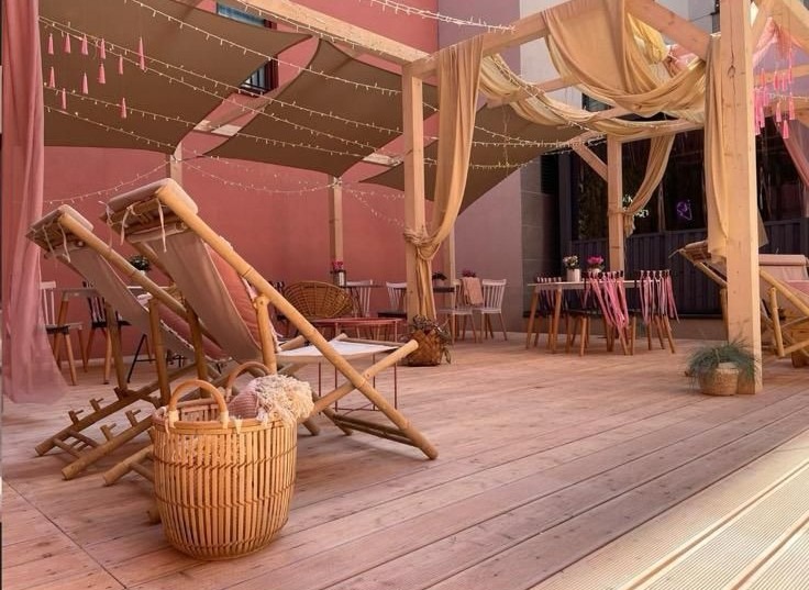 Drewniany taras z pergolą, leżakami i dekoracjami w odcieniach beżu i różu. Urokliwa aranżacja z oświetleniem i zasłonami tworzy relaksującą atmosferę.