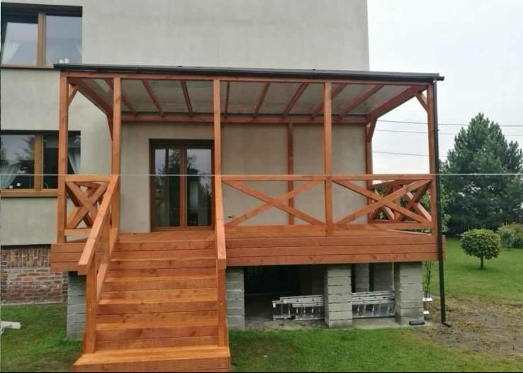 Drewniany taras z zadaszeniem przy domu, schody wejściowe, balustrada z krzyżowym wzorem, podmurówka z bloczków betonowych, widok na ogród.