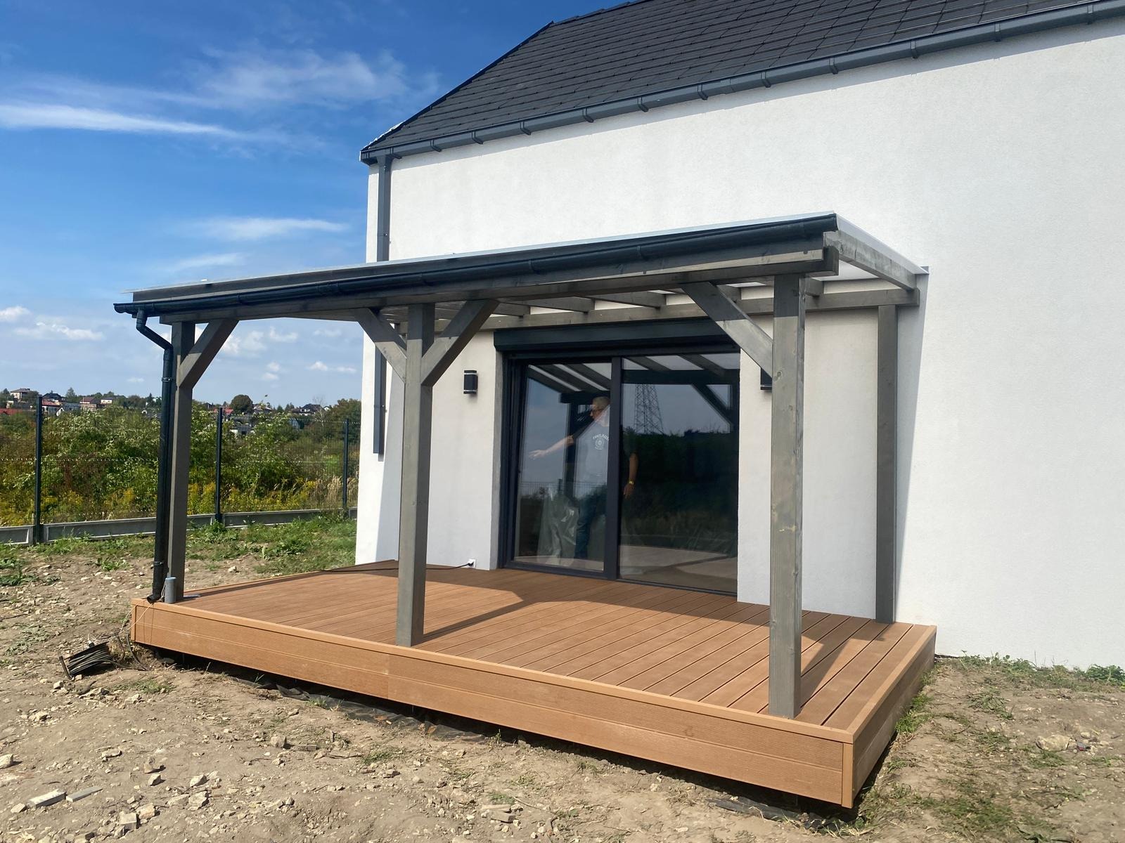 Drewniany taras kompozytowy z szarą pergolą przy białym domu. Widoczny fragment ogrodu i niebieskie niebo. Osoba w odbiciu szyby.