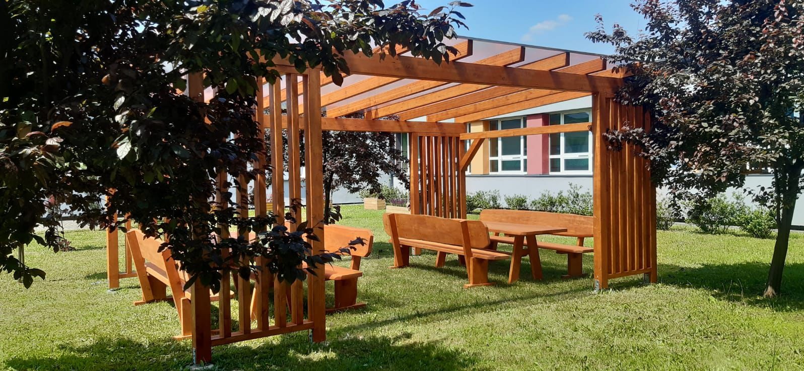 Drewniana pergola z zadaszeniem i kompletem mebli ogrodowych (stół i ławki) na trawniku. W tle budynek z oknami. Ujęcie zza drzewa.