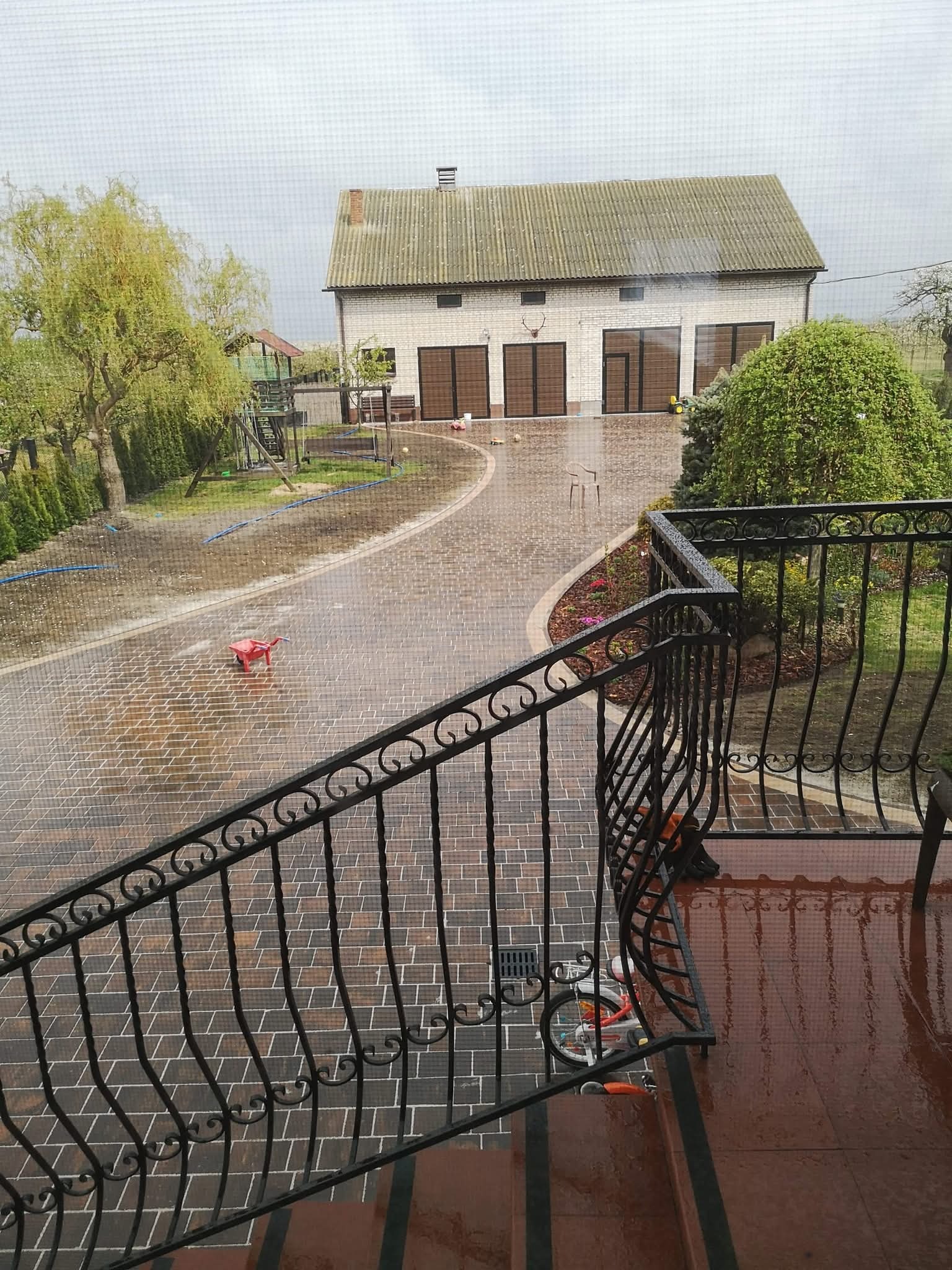 Deszczowy dzień na posesji z kostką brukową, widok z góry przez balustradę. W tle budynek gospodarczy i plac zabaw.