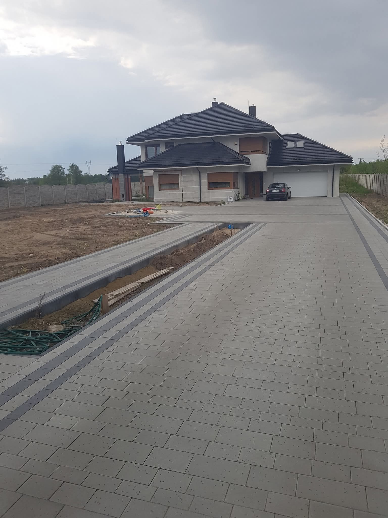 Nowoczesny podjazd z kostki brukowej prowadzący do domu z garażem, z widocznym przygotowanym miejscem pod trawnik i elementami instalacji nawadniającej.