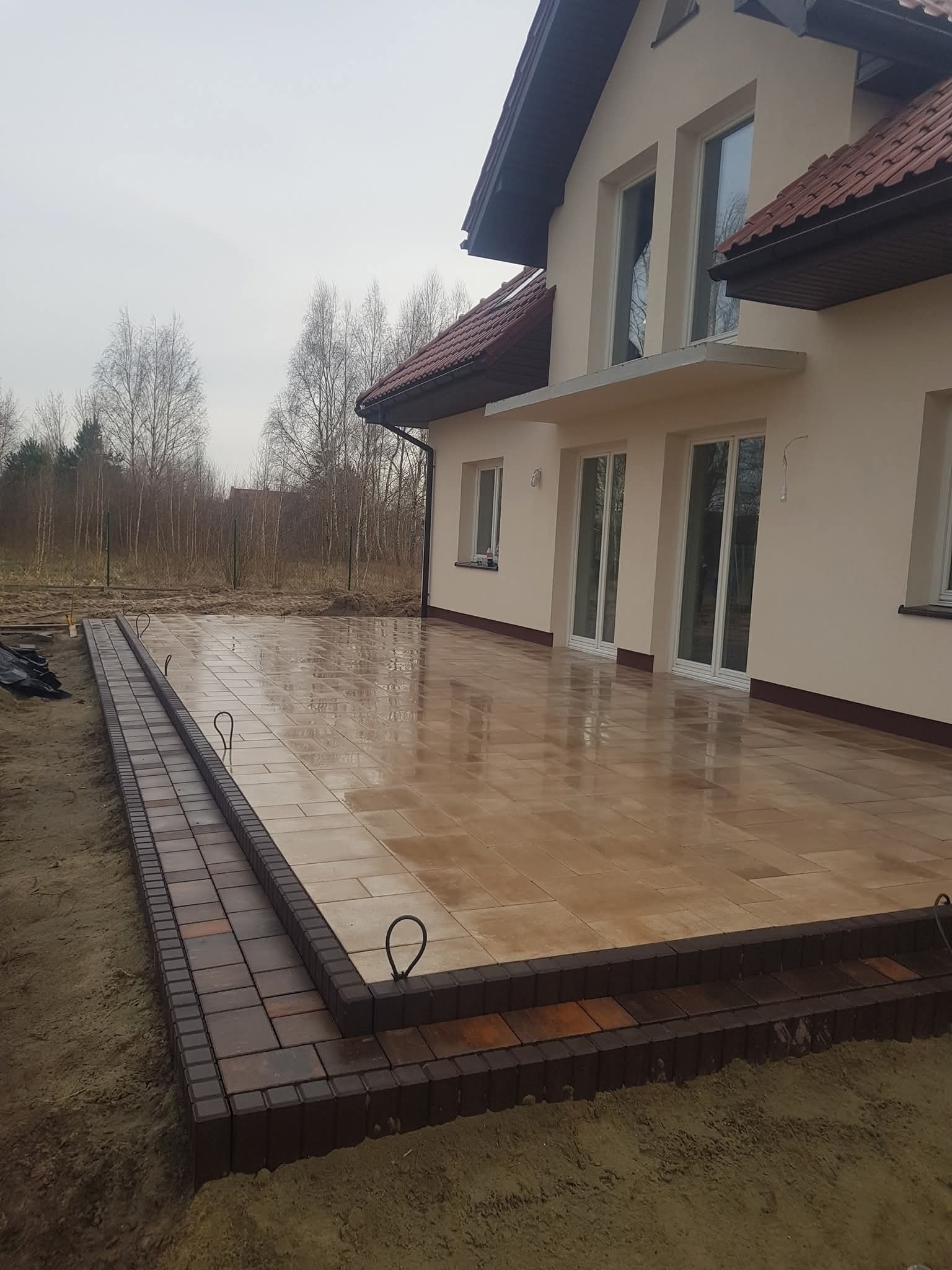 Ułożona, beżowa kostka brukowa na tarasie domu jednorodzinnego, z obrzeżem z ciemnobrązowej kostki. Widoczne metalowe uchwyty montażowe. Tło: drzewa bez liści.