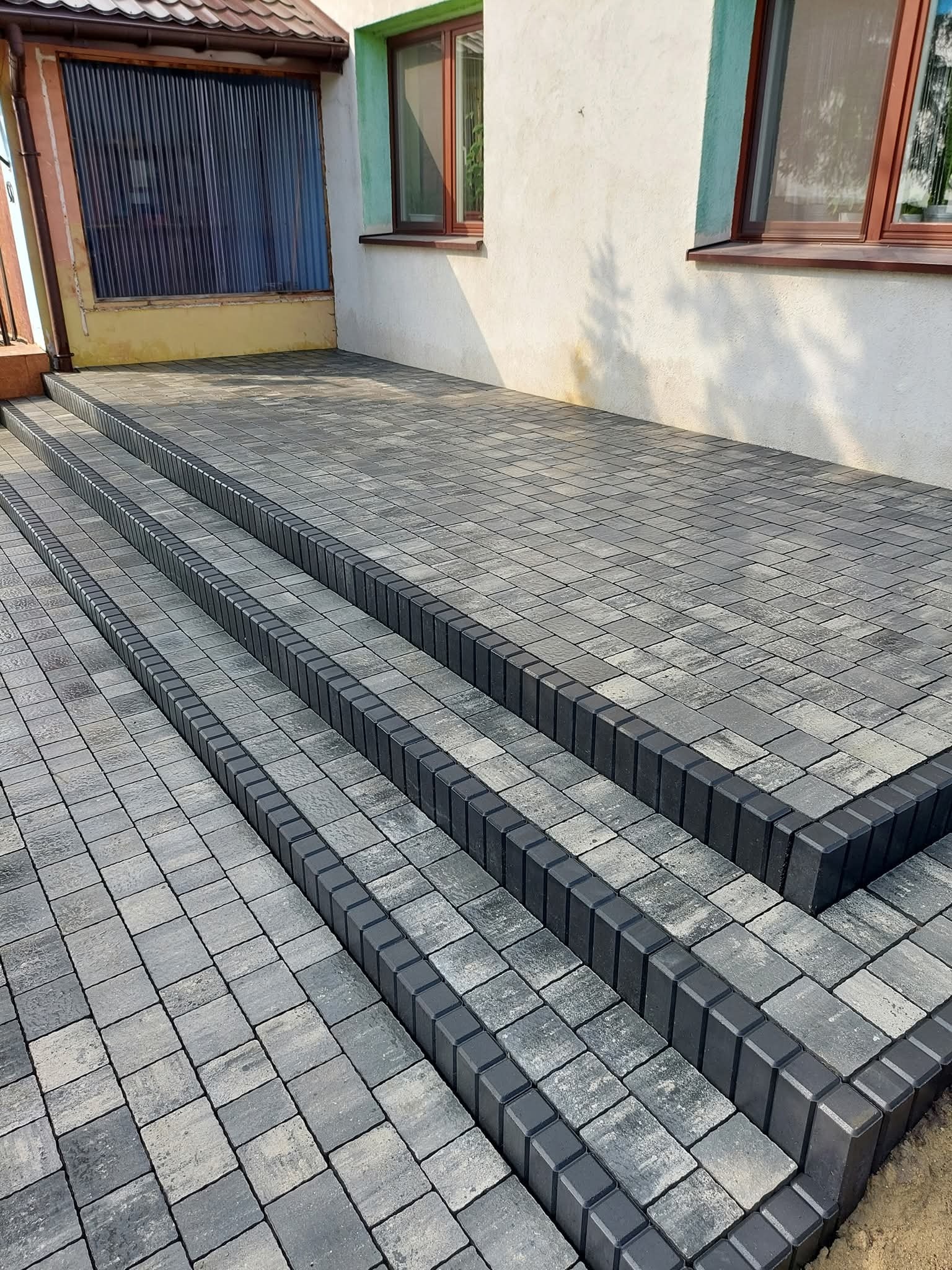Schody z kostki brukowej w odcieniach szarości, z ciemniejszym obramowaniem, prowadzące do budynku z oknami. Widok z dołu, podkreślający regularność wzoru.