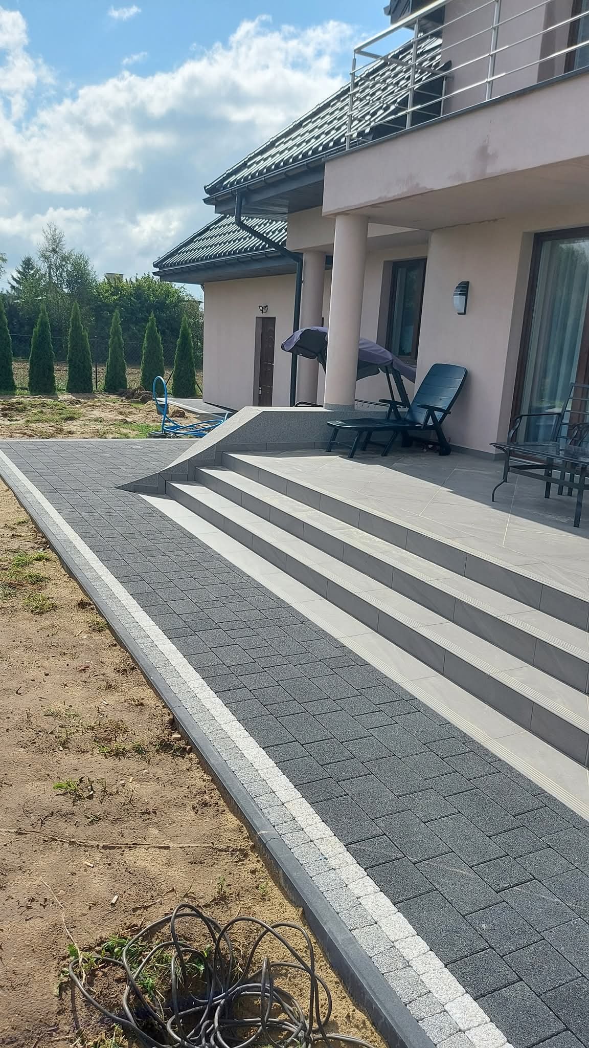 Ułożona szara kostka brukowa z białym pasem obok schodów wejściowych do domu. Widoczne krzesła ogrodowe i drzewa w tle. Całość w słoneczny dzień.