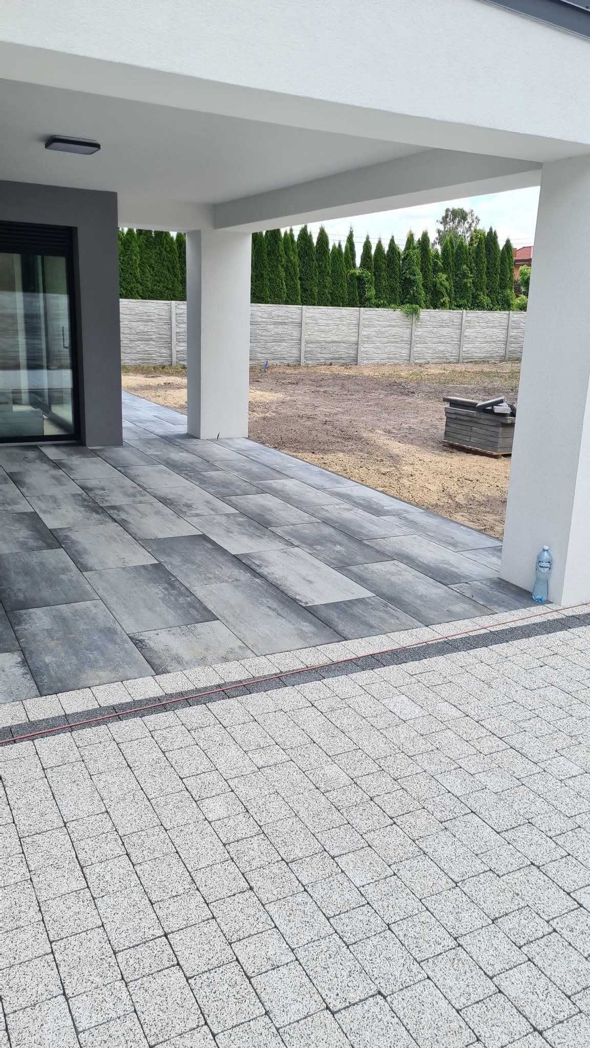 Nowoczesny taras z szarej kostki brukowej ułożonej pod zadaszeniem, z widokiem na ogrodzenie i przygotowany teren pod trawnik. Butelka wody przy kolumnie.