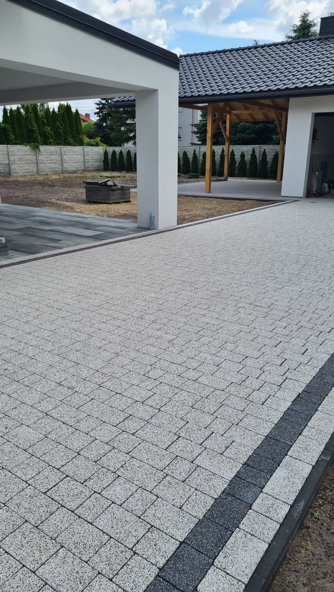 Jasnoszara kostka brukowa ułożona na podjeździe, z ciemnoszarym obramowaniem. W tle widoczny dom z drewnianą wiatą i uporządkowany teren.
