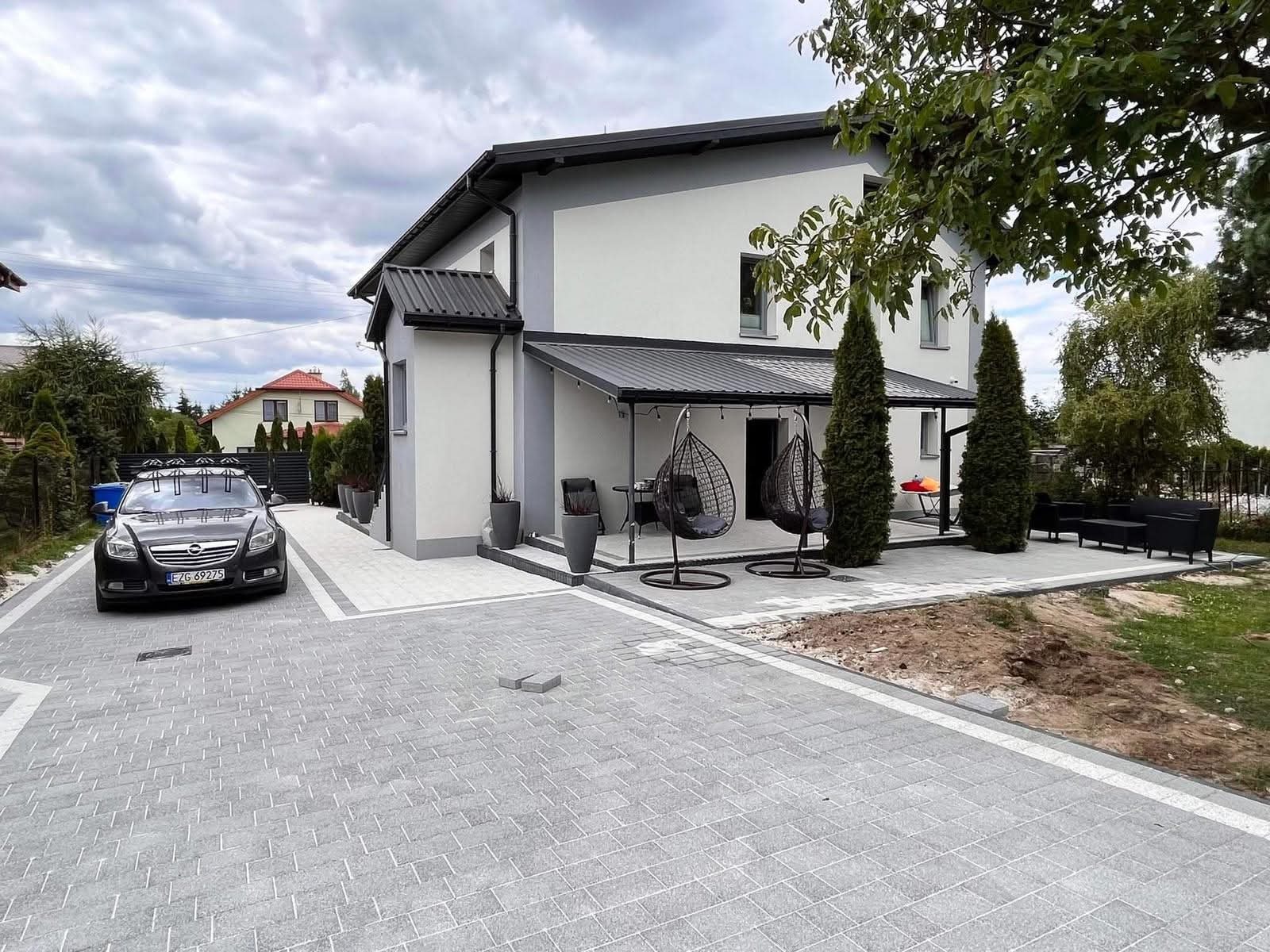 Nowoczesny dom z szarą kostką brukową na podjeździe i tarasie, uzupełniony o strefę relaksu z wiszącymi fotelami i eleganckimi donicami. Widoczny samochód osobowy.