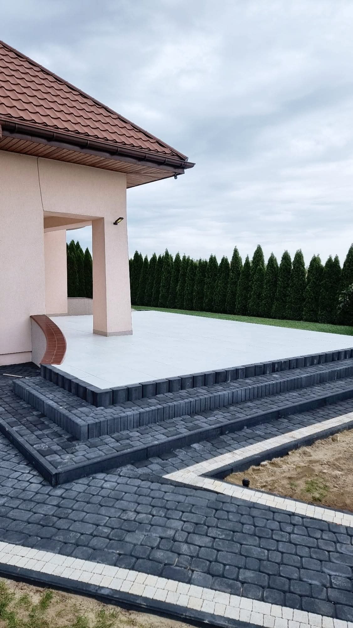 Nowoczesny taras z kostki brukowej w odcieniach szarości, z eleganckimi schodami i białymi akcentami, przylegający do jasnego budynku z brązowym dachem.