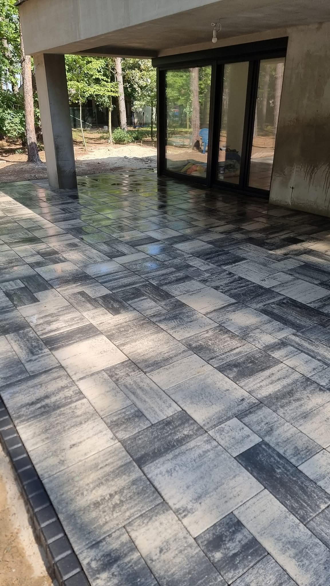 Nowoczesny taras z szarej kostki brukowej o nieregularnym wzorze, częściowo mokry, z widokiem na ogród przez przesuwne szklane drzwi.