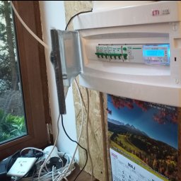 Dominik Borowski - Otwarta skrzynka elektryczna z bezpiecznikami i licznikiem, zamontowana na ścianie z płyty OSB, obok okna z widokiem na zewnątrz. Widoczne kable i kalendarz.