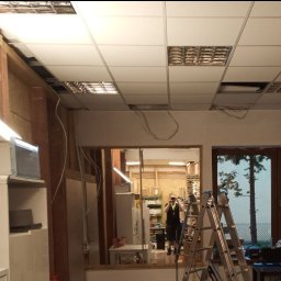 Dominik Borowski - Wnętrze w trakcie montażu sufitu podwieszanego z widocznymi przewodami elektrycznymi, drabina i częściowo widoczna osoba w tle.