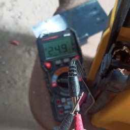 Dominik Borowski - Pomiar napięcia multimetrem cyfrowym, wartość 24.9 na wyświetlaczu, czarne i czerwone przewody pomiarowe w dłoni, żółta torba narzędziowa w tle.