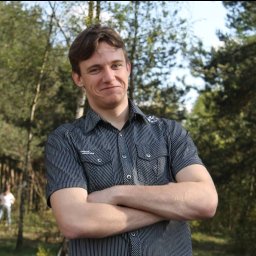 Dominik Borowski - Portret młodego mężczyzny w koszuli w paski, z założonymi rękami na tle zieleni drzew. Uśmiechnięty, z lekkim przymrużeniem oczu. Naturalne światło.