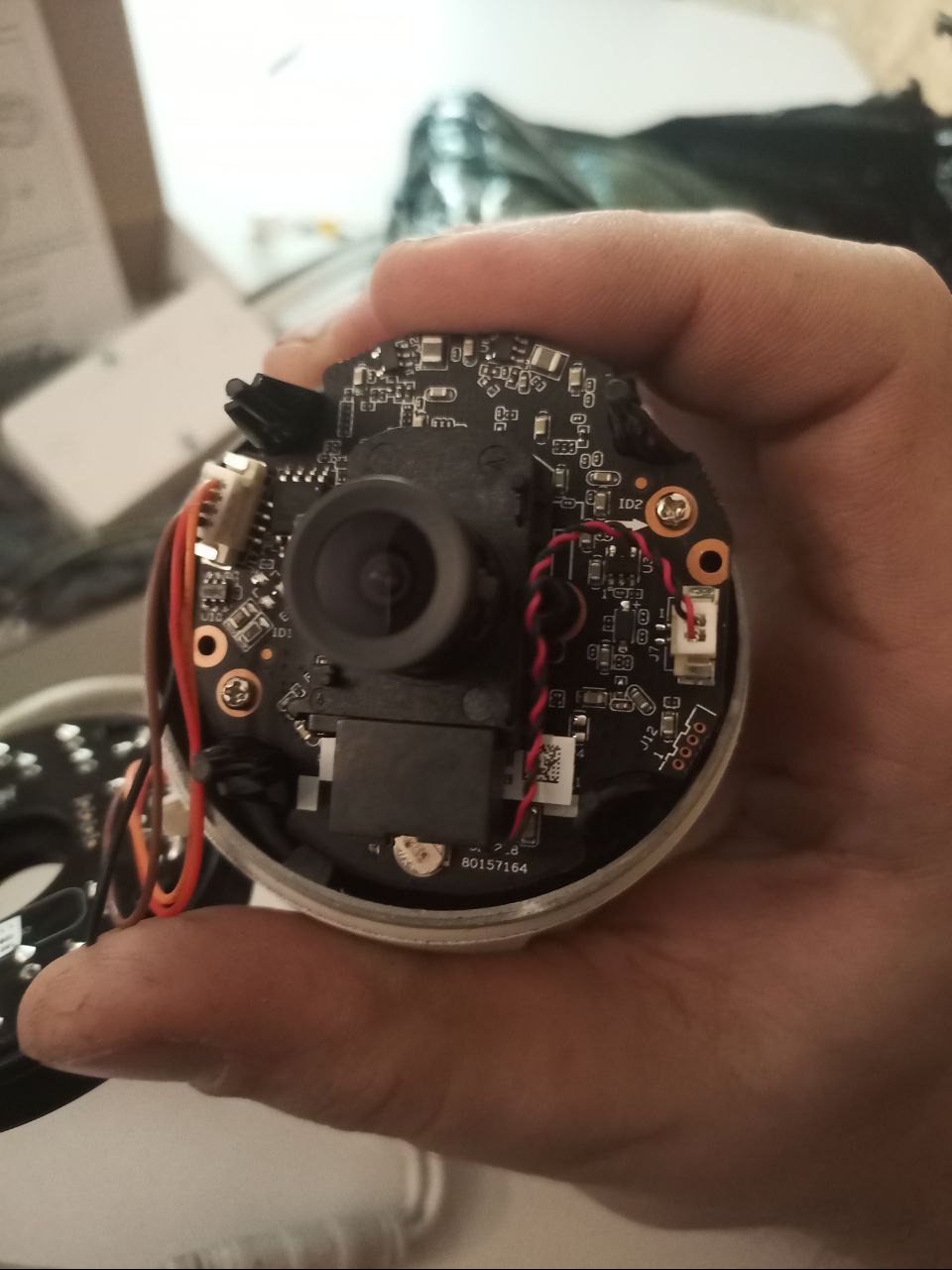 Otwarta obudowa kamery monitoringu z widoczną elektroniką, trzymana w dłoni. Widoczne przewody, obiektyw i płytka drukowana z elementami. Numer seryjny 80157164.