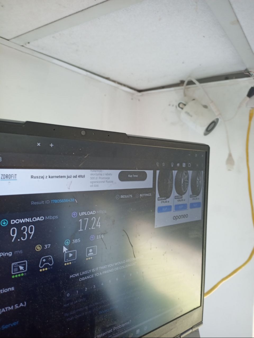Test prędkości internetu po instalacji kamery monitoringu. Laptop z uruchomionym testem, widoczna kamera na ścianie i kabel. Sprawdzanie jakości połączenia.