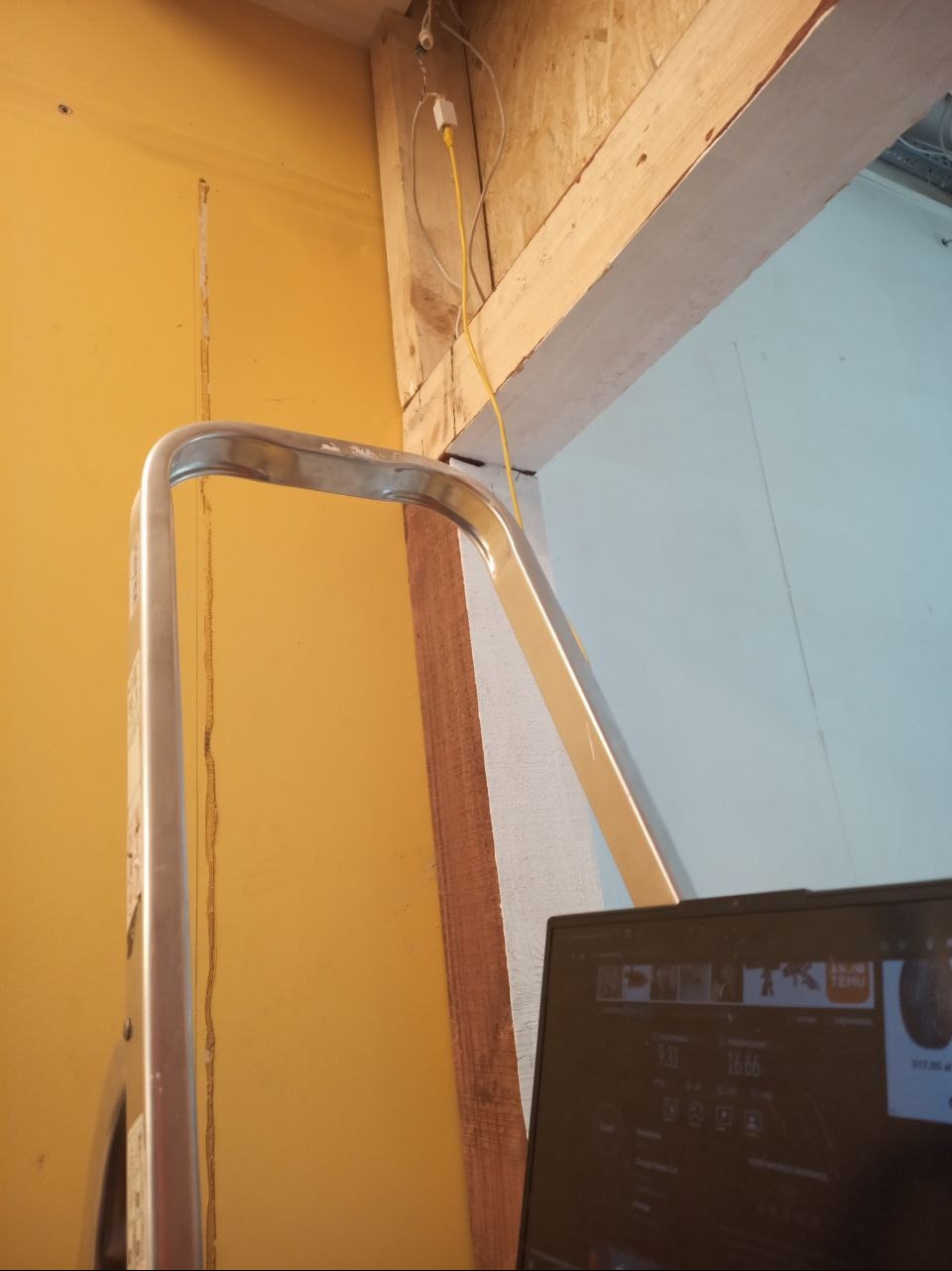 Żółta ściana z kablem i gniazdkiem, fragment drabiny aluminiowej i laptopa. Prace wykończeniowe wewnątrz budynku, widoczne elementy instalacji elektrycznej.