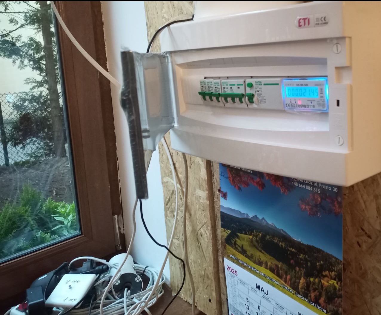 Otwarta skrzynka elektryczna z bezpiecznikami i licznikiem, zamontowana na ścianie z płyty OSB, obok okna z widokiem na zewnątrz. Widoczne kable i kalendarz.