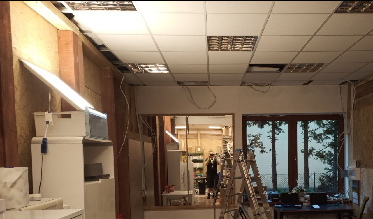 Wnętrze w trakcie montażu sufitu podwieszanego z widocznymi przewodami elektrycznymi, drabina i częściowo widoczna osoba w tle.