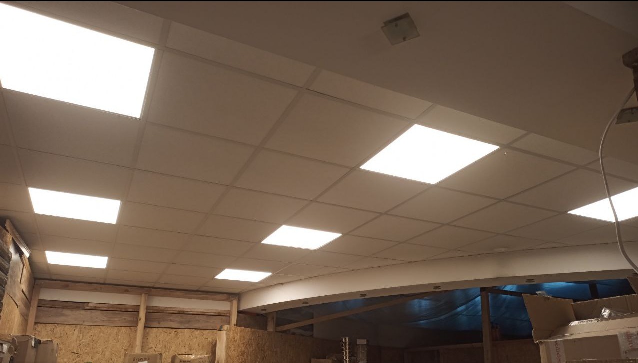 Sufit podwieszany z panelami LED w pomieszczeniu magazynowym. Widoczne elementy konstrukcyjne i okablowanie. Oświetlenie równomierne, jasne.