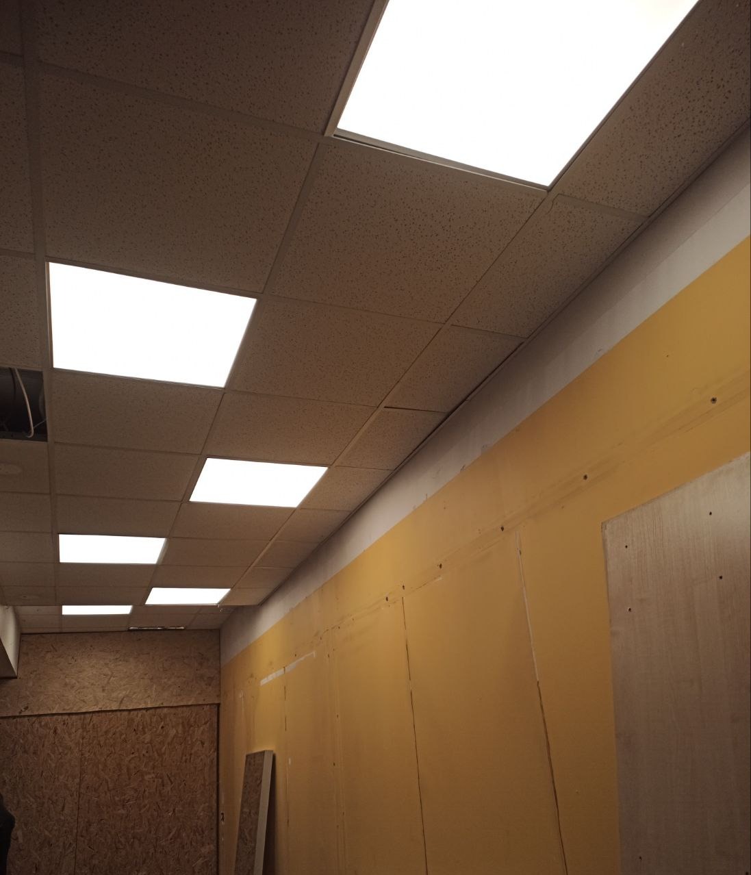 Kwadratowe panele LED w suficie podwieszanym, oświetlające żółtą ścianę w pomieszczeniu w trakcie remontu. Widoczne płyty OSB na ścianie po lewej stronie.