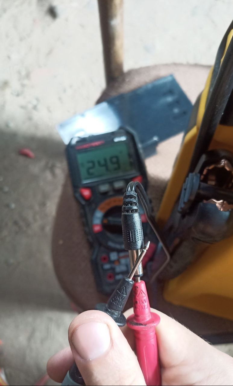 Pomiar napięcia multimetrem cyfrowym, wartość 24.9 na wyświetlaczu, czarne i czerwone przewody pomiarowe w dłoni, żółta torba narzędziowa w tle.