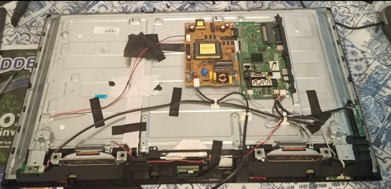 Otwarty telewizor LCD z widocznymi płytkami drukowanymi, kablami i głośnikami, zabezpieczonymi taśmą. Widok od tyłu, ukazujący wewnętrzną konstrukcję urządzenia.