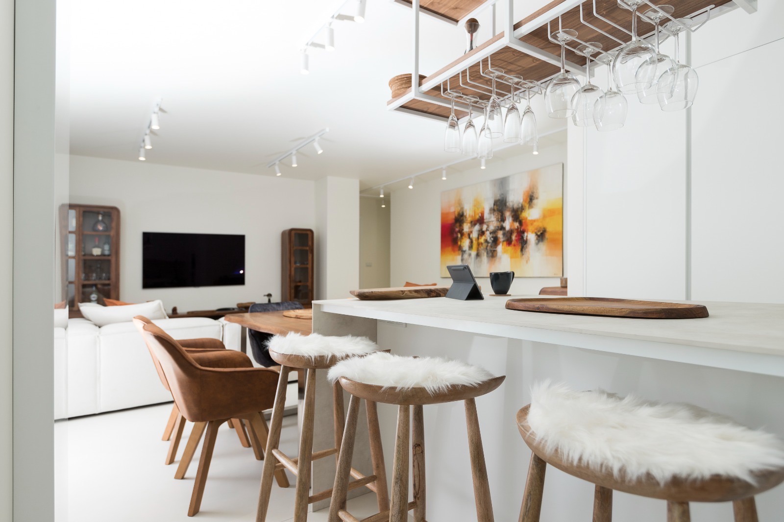 Jasne wnętrze z wyspą kuchenną, drewnianymi stołkami z białymi poduszkami, wiszącym regałem na kieliszki i abstrakcyjnym obrazem w tle. Nowoczesny design.