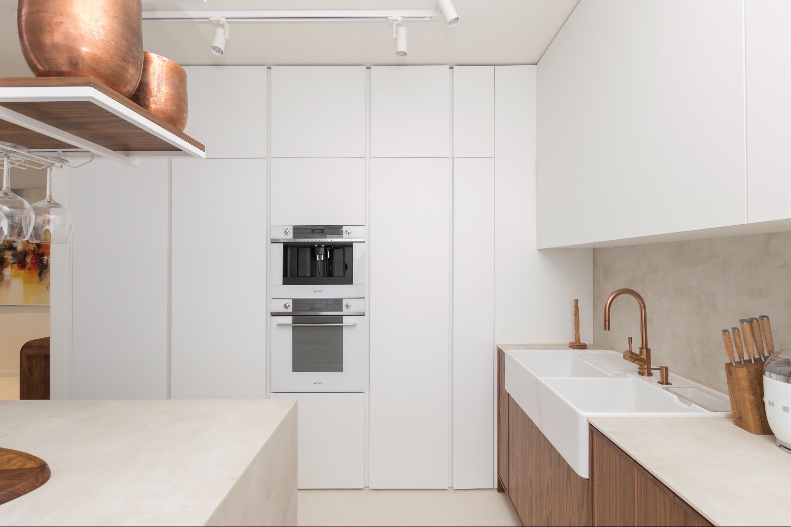 Minimalistyczna kuchnia: białe szafki, wbudowany sprzęt Smeg, miedziana armatura i dekoracje. Jasne, nowoczesne wnętrze z wyspą i widocznymi kieliszkami.